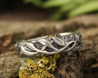 May include: A silver ring with a detailed design of leaves and branches. The ring is resting on a piece of wood with green moss and yellow lichen.