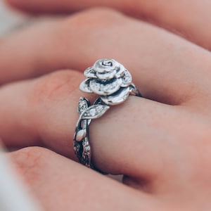 Peut inclure: Une bague en argent avec un motif de rose et des pierres claires. La bague est portée sur un doigt, avec un livre flou en arrière-plan.