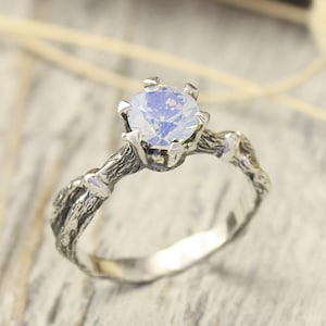 May include: A silver ring with a white opal gemstone set in a twig-like band. The ring is on a light wood background.