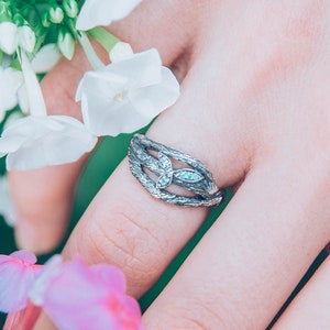 Könnte beinhalten: Ein silberner Ring mit einem filigranen Design, das einen kleinen, klaren Stein zeigt. Der Ring wird an einer Hand getragen, die einen Strauß weißer und rosa Blumen hält.