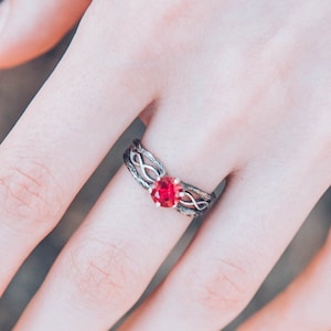 May include: A silver ring with an intricate, interwoven design. The ring features a vibrant, oval-shaped red gemstone at its centre. The ring is displayed on a hand, with a blurred natural background.