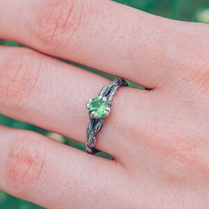 Op de afbeelding: Een zilveren ring met een groene edelsteen gezet in een delicate, gedraaide band. De ring wordt gedragen op een hand tegen een wazige achtergrond van groen gebladerte.