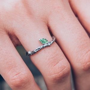 Puede incluir: Un anillo de plata con un diseño de rama y una hoja de piedra preciosa verde. El anillo se lleva en un dedo.