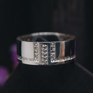 May include: A silver ring with a geometric design. The ring features a rectangular shape with a line of small, clear stones. The ring is set against a dark background, highlighting its polished surface. The ring is a piece of jewelry.