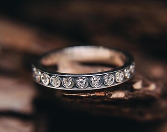 Puede incluir: Un anillo de plata con una banda de pequeñas piedras preciosas transparentes. El anillo se encuentra sobre una superficie de madera oscura y texturizada, creando un contraste que resalta los detalles del anillo.