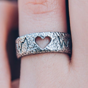 May include: A silver ring with a heart-shaped cutout and a textured surface. The ring features a heartbeat design around the band. The ring is worn on a finger.