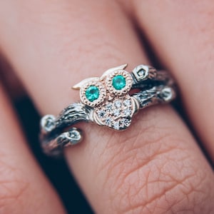 Könnte beinhalten: Ein Ring mit einem Eulenmotiv, zwei grünen runden Steinen als Augen und kleinen klaren Steinen auf der Brust. Die Ringschiene ist wie ein Baumzweig strukturiert, mit einem dunklen Metallfinish und roségoldenen Akzenten am Kopf der Eule.