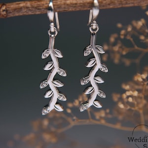 May include: Silver dangle earrings with a delicate vine design and small clear stones. The earrings are hanging from a branch.