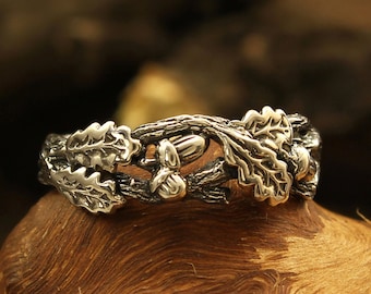 May include: A silver ring with an oak leaf and acorn design. The ring is on a wooden surface.