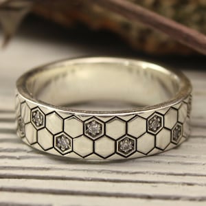 May include: A silver wedding band with a honeycomb pattern and small clear stones. The band is on a light wood surface.