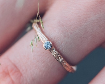 May include: A delicate gold band with a small, clear gemstone set in the center. The ring is worn on a hand with a textured, organic design.