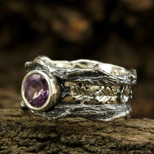 May include: A silver ring with a purple amethyst gemstone set in a textured band that resembles a tree branch. The ring is on a wooden surface.