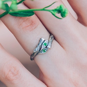 May include: A silver ring with a nature-inspired design featuring a branch-like band and small green gemstones. The ring is worn on a finger, with a sprig of white flowers in the background. The ring's design is intricate and detailed.
