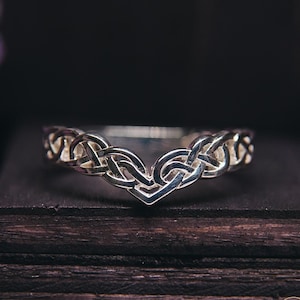 Puede incluir: Un anillo plateado con un diseño de nudo celta. El anillo tiene un centro en forma de V y un intrincado trabajo de nudos a lo largo de la banda. Se muestra sobre una superficie de madera oscura, con flores moradas en el fondo.