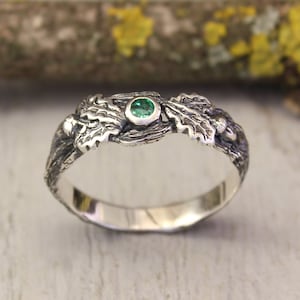 Può includere: Un anello d'argento con una pietra preziosa verde incastonata al centro, circondato da un design dettagliato di foglie e ghiande.