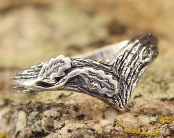 May include: A silver ring with a detailed design of a branch with leaves. The ring is set on a textured rock with green moss.