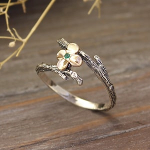 Może przedstawiać: Srebrny pierścionek z kwiatowym wzorem. Pierścionek ma opaskę w kształcie gałęzi i kwiat z zielonym kamieniem pośrodku i złotymi płatkami. Pierścionek jest umieszczony na drewnianym tle.
