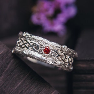 Op de afbeelding: Een zilveren trouwring met een Keltisch knoopontwerp en een rode edelsteen. De ring is getextureerd om op boomschors te lijken.