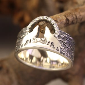 May include: A silver ring with a textured band and two wolf silhouettes facing each other. The wolves are outlined with a row of small clear stones.