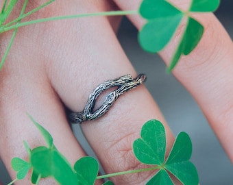 May include: A silver ring with a unique design resembling intertwined branches. The ring is worn on a finger with green clover leaves in the background.