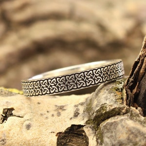 May include: A silver wedding band with a Celtic knot design. The band is sitting on a piece of weathered wood.