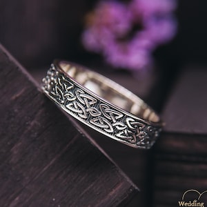 May include: A silver wedding band with a Celtic knot design. The band is on a dark wooden surface.