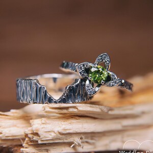 Puede incluir: Dos anillos de plata. Uno tiene una banda texturizada con un diseño de montaña. El otro presenta un engaste en forma de flor con una piedra preciosa verde en el centro y pequeñas piedras circundantes. Los anillos se exhiben sobre un trozo de madera.