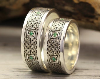 May include: Two silver wedding bands with a Celtic knot design and green gemstones. The rings are on a wooden surface.