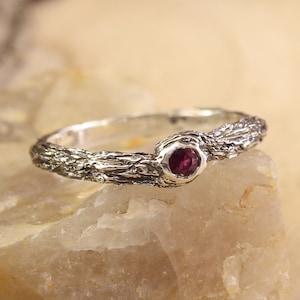 Peut inclure: Une bague en argent avec une bande texturée ressemblant à de l'écorce d'arbre et une petite pierre précieuse ronde rose foncé.