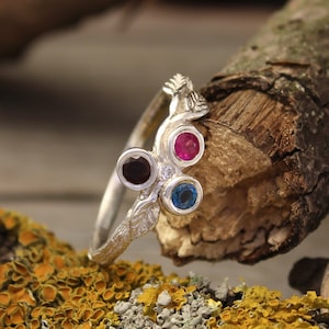 Può includere: Un anello d'argento con un delicato design a ramo e tre pietre preziose: una rosso scuro, una rosa e una blu.