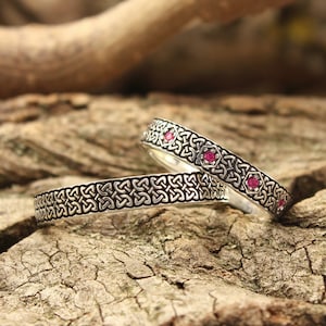 May include: Two silver Celtic knot wedding bands with pink gemstones. The bands are on a wooden surface.