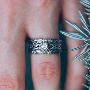 May include: A silver wedding band with a Celtic knot design and a small diamond in the centre. The ring is worn on a finger against a green background.