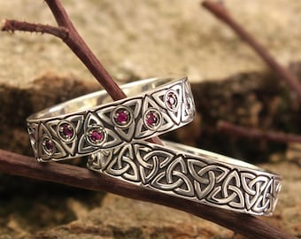 May include: Two silver Celtic knot wedding bands with pink gemstones. The bands are intertwined and resting on a piece of tree bark.