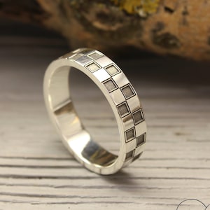May include: A silver wedding band with a checkered pattern. The band is on a white wooden surface.