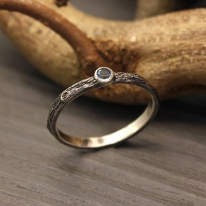 Könnte beinhalten: Ein Silberring mit einer strukturierten, rindenartigen Oberfläche und einem einzelnen, kleinen, runden blauen Edelstein in einer Fassung. Das Band hat ein oxidiertes Finish, das die komplexen, organischen Details der Metallarbeit rundherum betont.