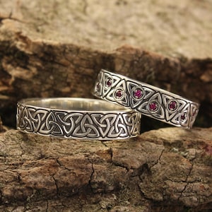 May include: Two silver wedding rings with Celtic knot designs. One ring features a continuous knot pattern, while the other has triangular patterns with red gemstones. The rings are displayed on a piece of textured wood.