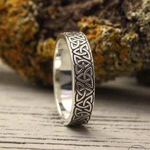 May include: A silver wedding band with a Celtic knot design. The band is on a wooden surface.