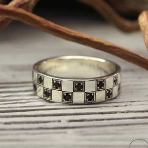 May include: A silver band with a checkered pattern of black square stones. The ring is on a wooden surface.