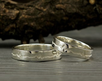 May include: Two silver wedding bands with a heartbeat design. The bands are on a wooden surface.