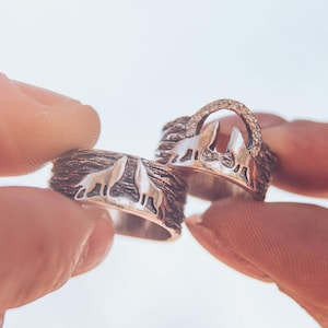 May include: Two silver rings with a wolf design. One ring features a band of small diamonds. The rings are held up against a bright background. The rings have a textured band and a wolf silhouette.
