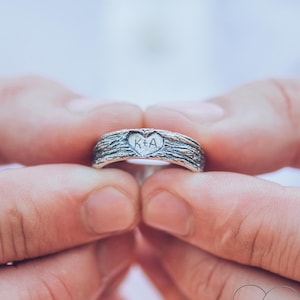 Op de afbeelding: Een zilveren trouwring met een hartvormig ontwerp en de initialen "K+A" gegraveerd aan de binnenkant. De ring heeft een getextureerd patroon dat lijkt op boomschors.