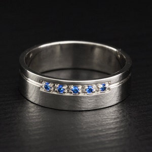 Może przedstawiać: Złota obrączka ślubna z rzędem niebieskich kamieni.