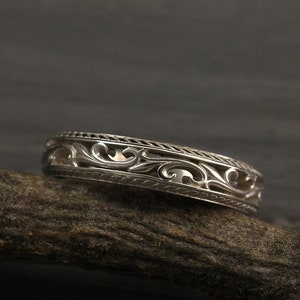 May include: A silver wedding band featuring a delicate, intricate floral design. The band is resting on a piece of driftwood.