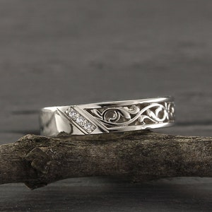 Peut inclure: Une alliance en argent avec un design délicat et complexe, ornée d'un motif de vigne et de petits diamants. La bague est posée sur une branche.