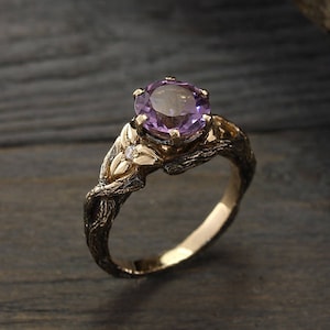 May include: A gold engagement ring with a purple amethyst gemstone and a leaf design. The ring is set on a wooden surface.