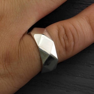 May include: A silver ring with a geometric design. The ring is worn on a person's finger.