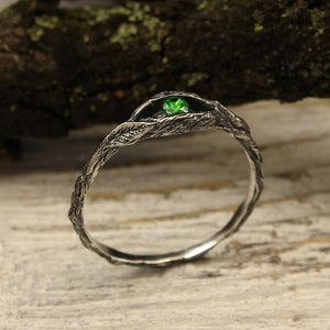 Op de afbeelding: Een zilveren ring met een getextureerd, takachtig ontwerp, met een kleine, ronde, heldergroene edelsteen. De ring staat tegen een rustieke achtergrond met een houten tak en een lichtgekleurd oppervlak. Het is een sieraad.