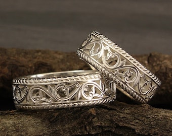 May include: Two silver wedding bands with intricate, ornate designs. The bands are stacked on top of each other and are resting on a piece of wood.