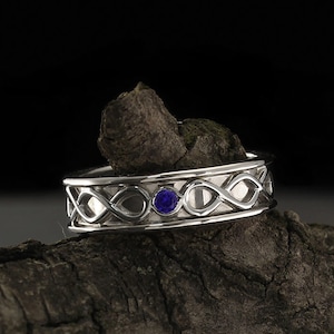 Könnte beinhalten: Ein silberner Ehering mit einem keltischen Knotendesign und einem einzelnen blauen Edelstein.