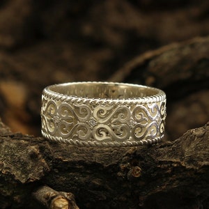 May include: A silver wedding band with a delicate, intricate design featuring swirls and small diamonds. The band is resting on a piece of weathered wood.